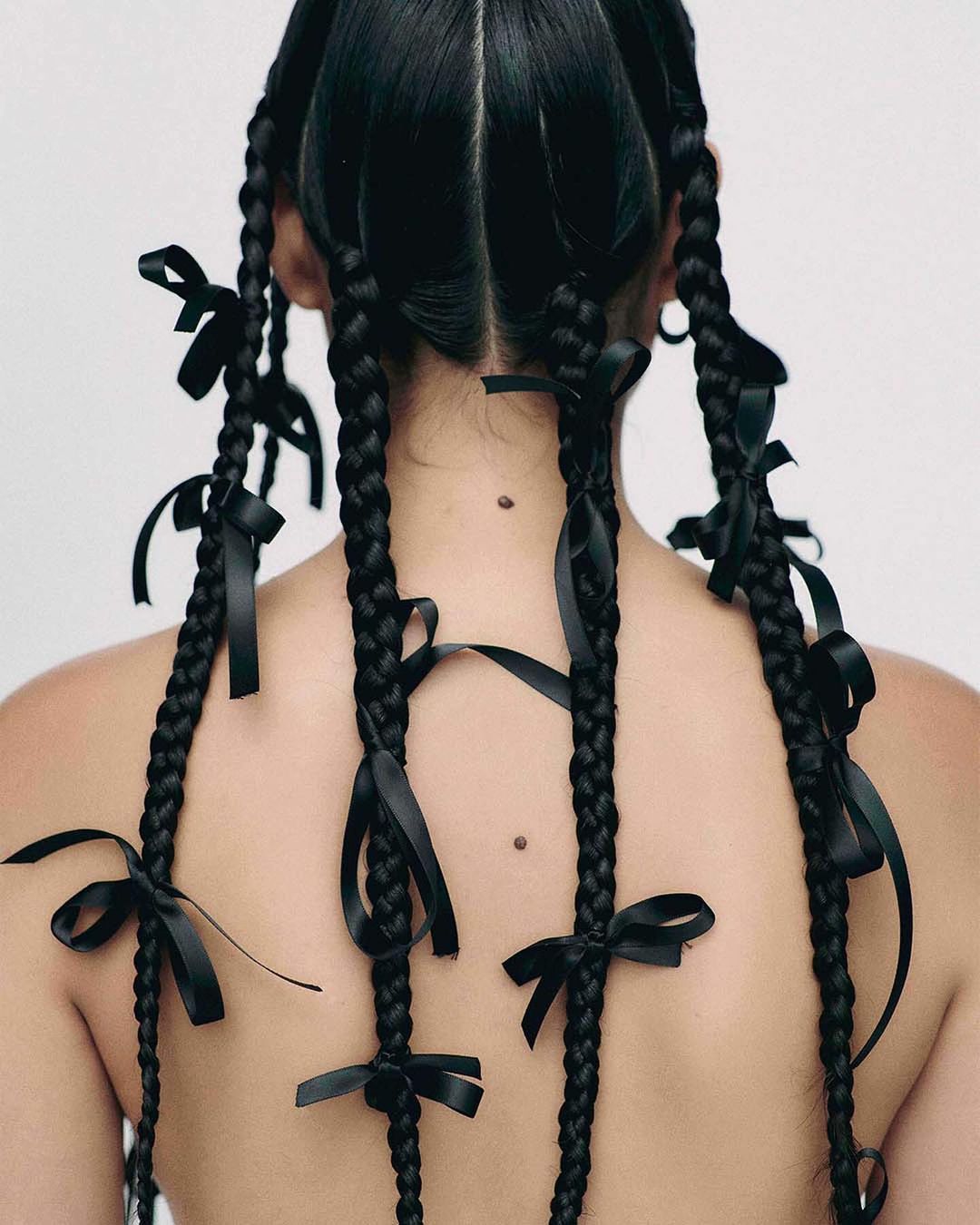 Rear view of a person with black braided hair adorned with tied black satin ribbons against a plain white backdrop in a studio setting.