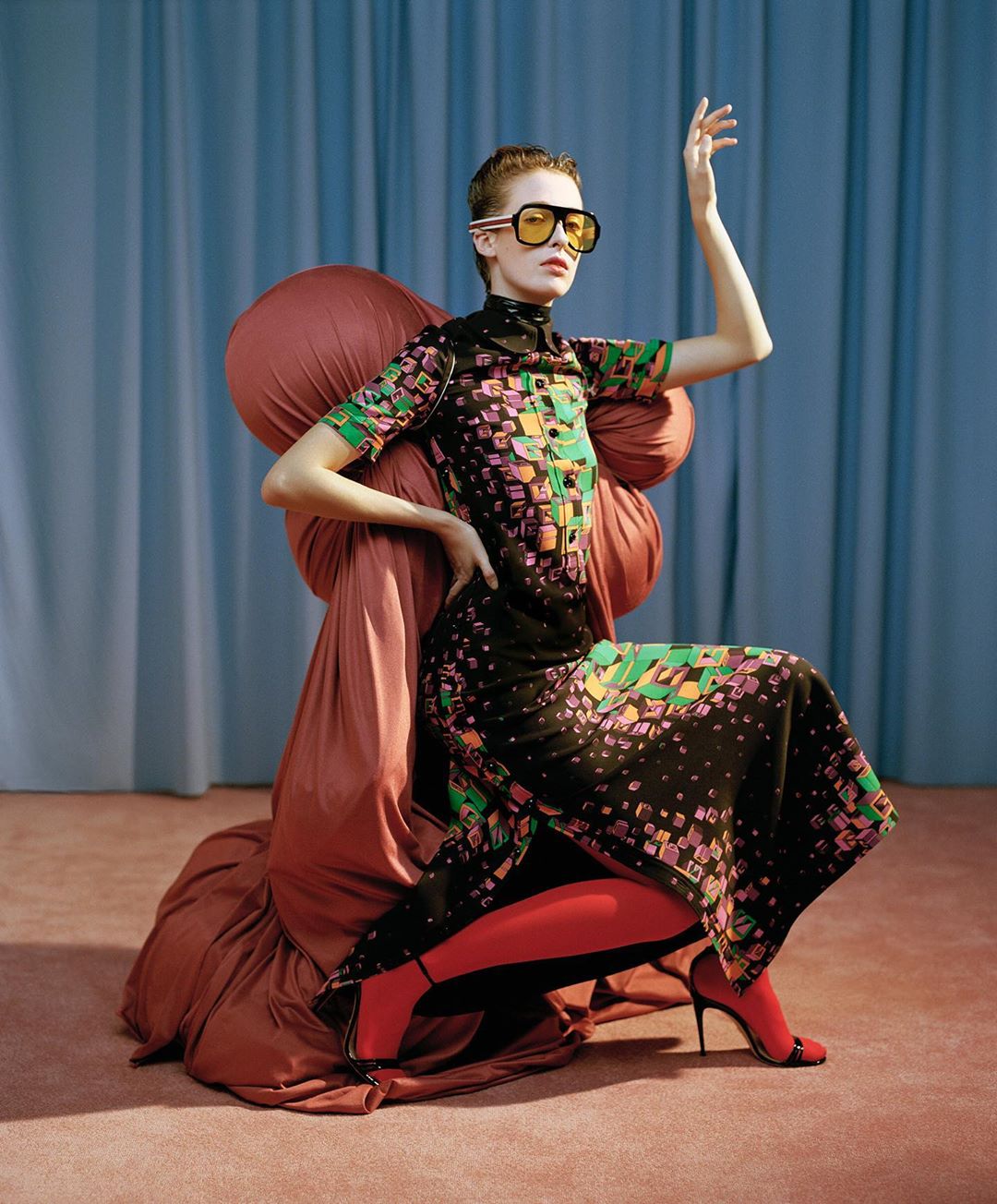 Fashion model poses in a black geometric print dress, red tights, and yellow sunglasses, seated on a draped peach-colored chair against blue curtains.