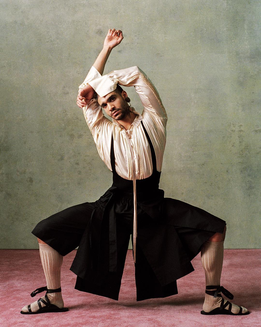 Person in a squat stance wearing a white ruffled shirt, black pants, and strappy sandals against a muted green and pink background.
