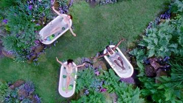 Three women relaxing in bathtubs placed in a lush green garden, viewed from above, creating a surreal and peaceful scene.