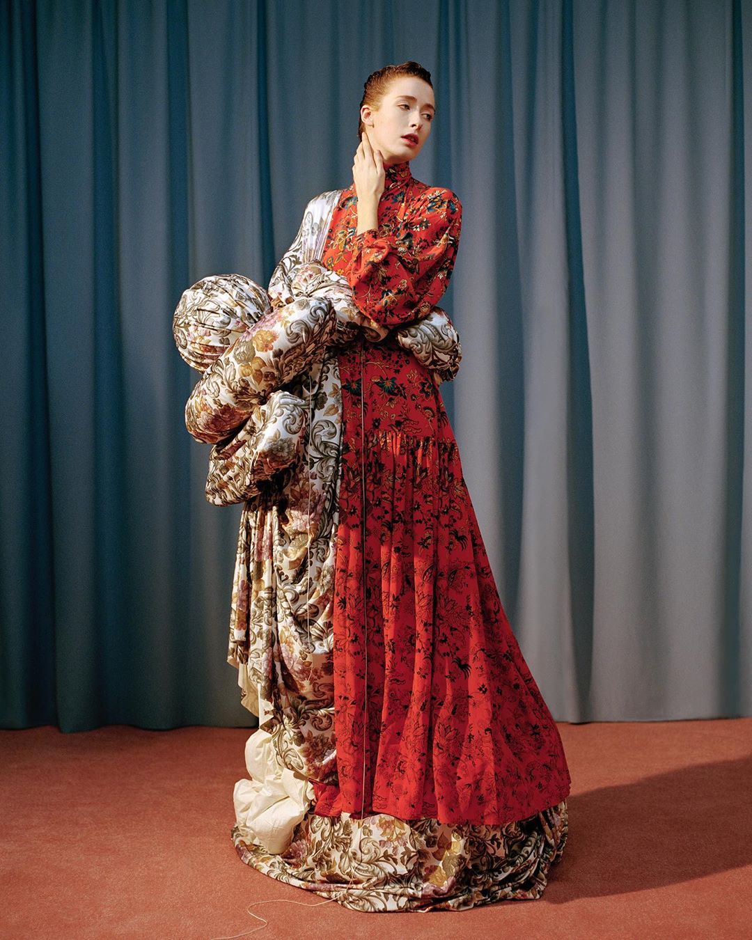 A fashion shot of a slender woman in a red floral dress posing next to a draped fabric statue against a blue curtain backdrop.