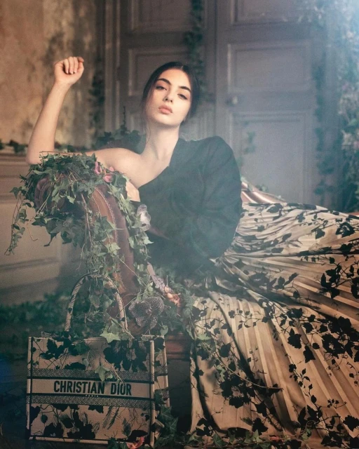 Elegant woman in couture poses with designer handbag in a lush interior with floral accents, styled for a high-fashion editorial.