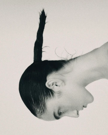 Profile portrait of a person with dark slicked-back hair styled into a pointed vertical ponytail against a light background.