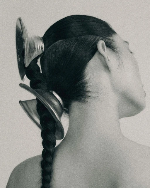 Monochromatic portrait of a woman with dark hair styled in a braid and adorned with metallic, concave hair accessories.