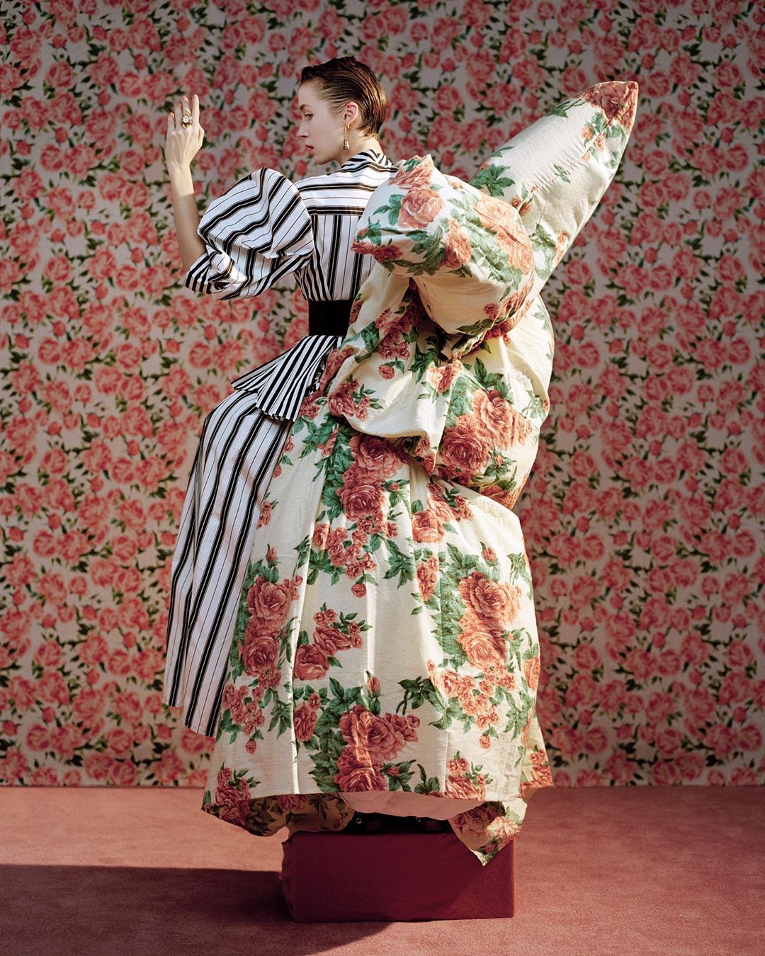 Back view of a fashion model posing on a box in a studio with floral wallpaper wearing a striped jacket and floral-print dress.
