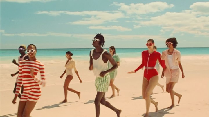 Group of women run on a beach wearing vintage-style swimsuits and sunglasses, with the ocean and sky in the background.