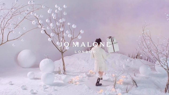 A woman walks through a snow-covered landscape with trees decorated with white spheres, carrying stacked white gift boxes with black ribbon.