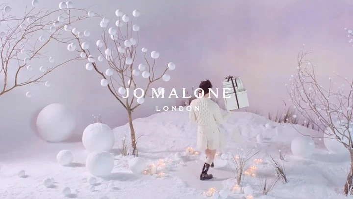 A woman walks through a snow-covered landscape with trees decorated with white spheres, carrying stacked white gift boxes with black ribbon.