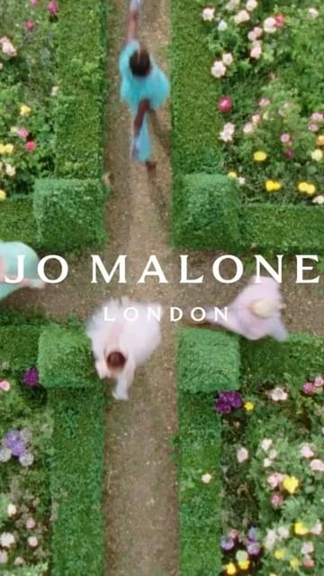 Overhead aerial view of models walking along a hedge-lined garden path with Jo Malone London text overlay in a bright, summery scene.
