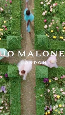 Overhead aerial view of models walking along a hedge-lined garden path with Jo Malone London text overlay in a bright, summery scene.