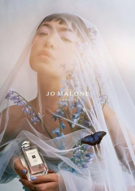 Cosmetic ad: Woman draped in sheer fabric holds a perfume bottle surrounded by flowers and a butterfly, showcasing a beauty product.
