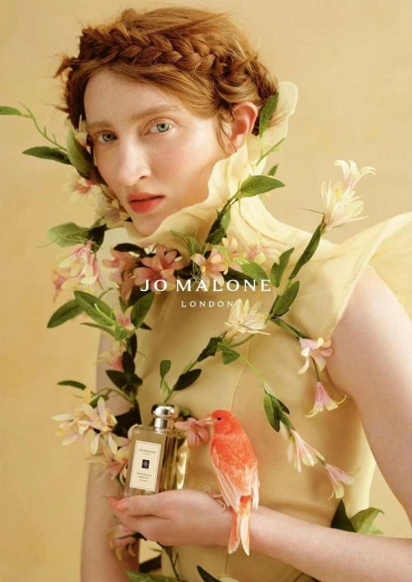 Elegant beauty portrait of a red-haired woman wearing floral decorations, holding perfume with a bird, on a yellow background.