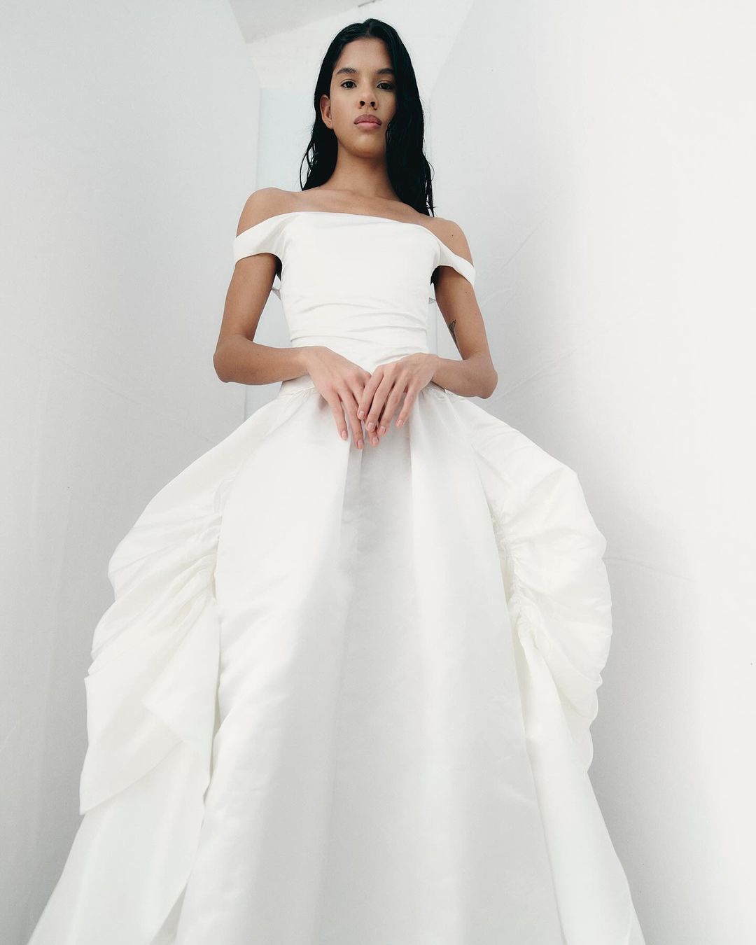 Woman in an off-the-shoulder white ballgown stands against a white wall, hands clasped, looking at the camera.