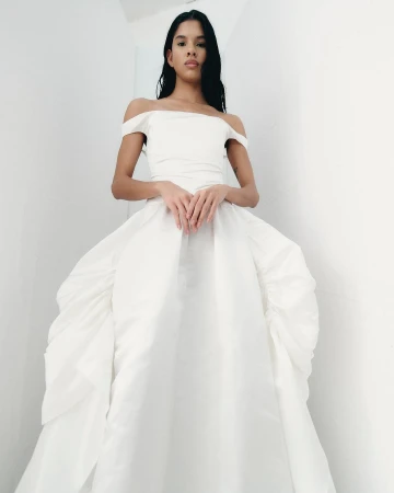 Woman in an off-the-shoulder white ballgown stands against a white wall, hands clasped, looking at the camera.