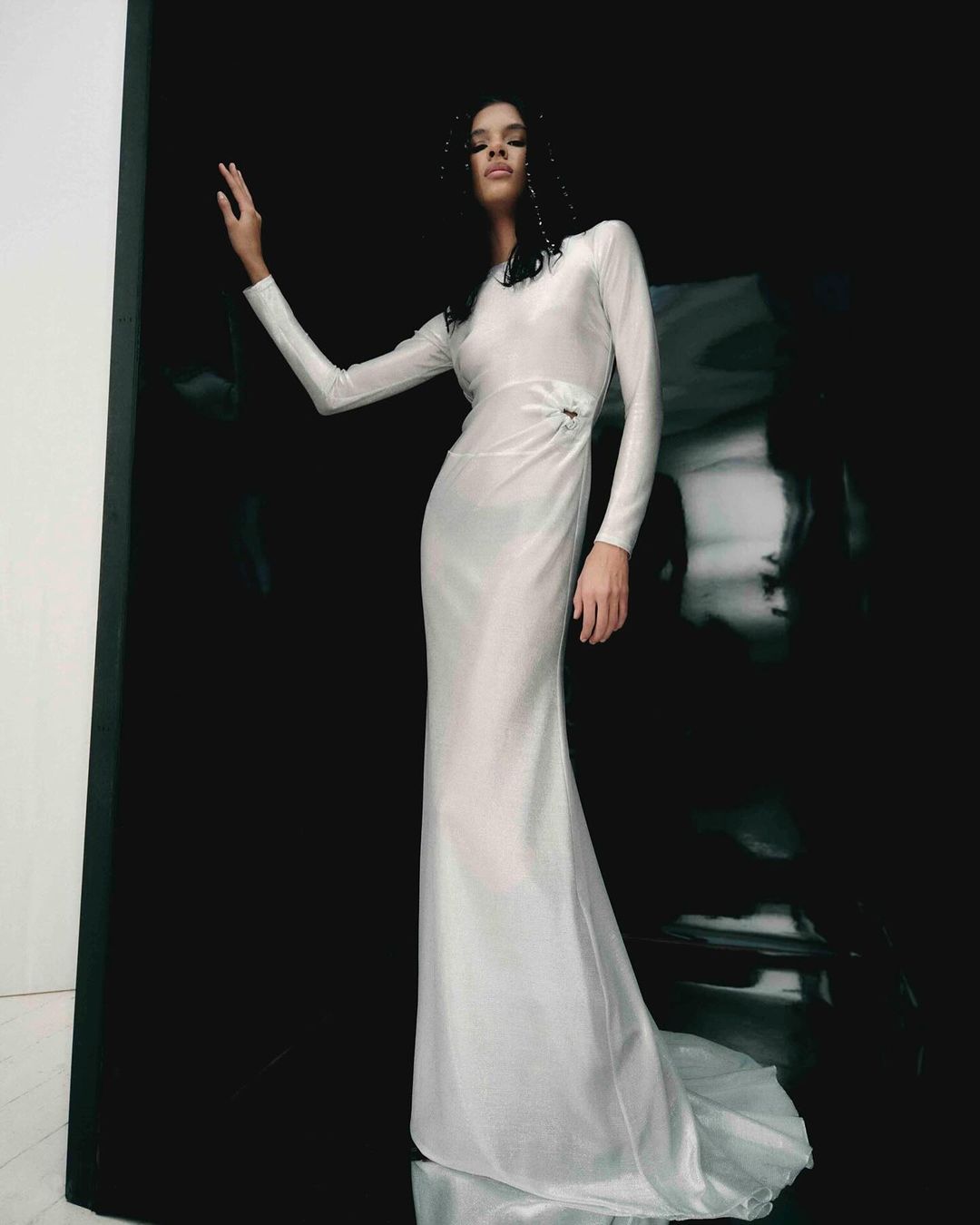 Elegant adult female poses in a long-sleeved, floor-length white dress against a striking black background in a fashion photoshoot.