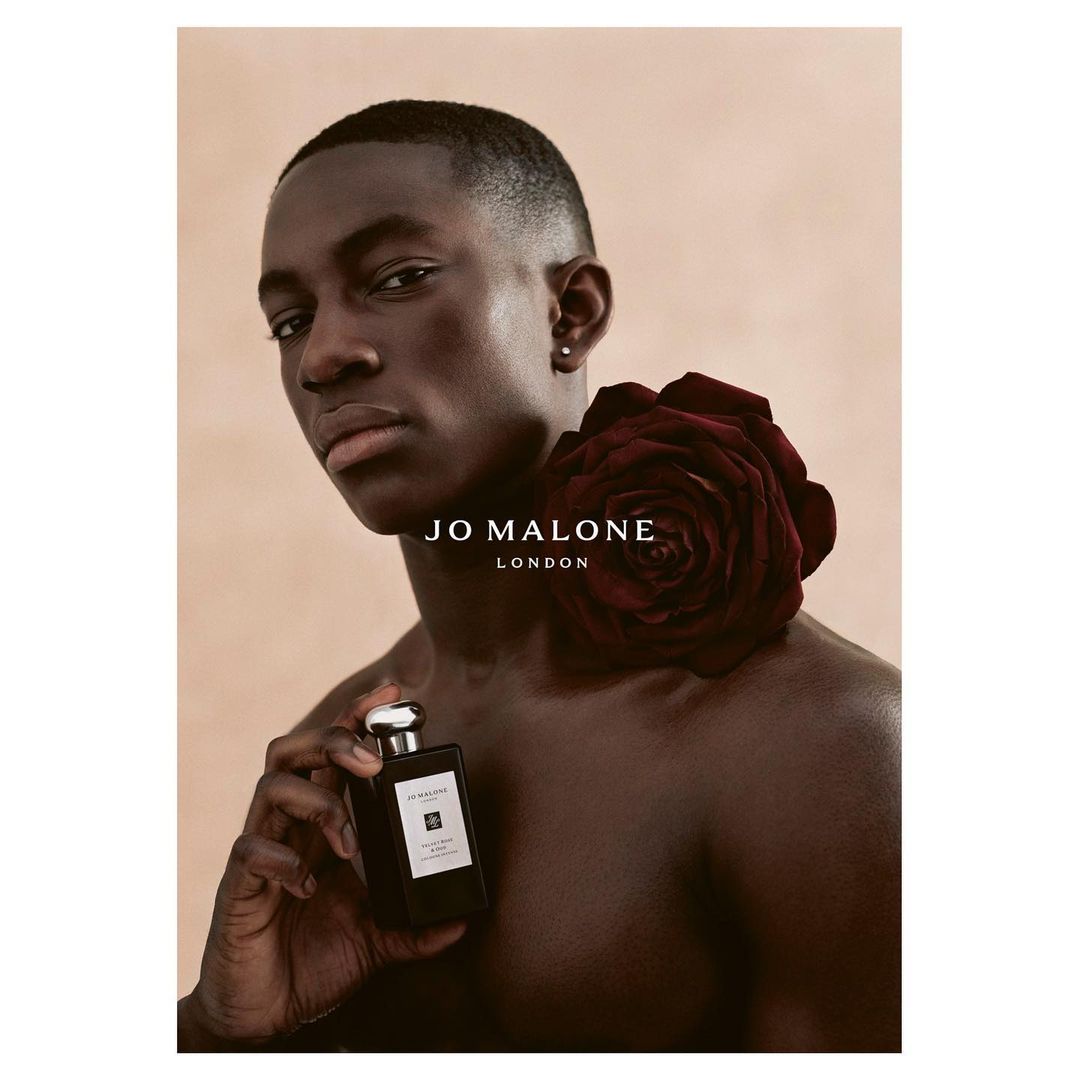 Dark-skinned male portrait with a dark rose on his shoulder, holding a perfume bottle, and the Jo Malone London logo.