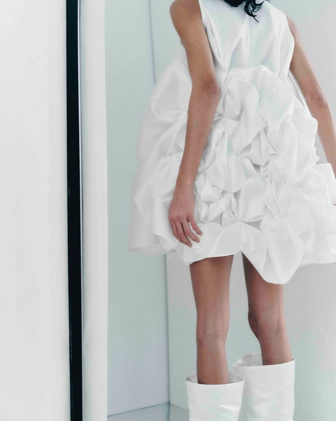 Fashion model wears a short white dress with layers of ruffles and white boots in a minimalist white photo studio setting.