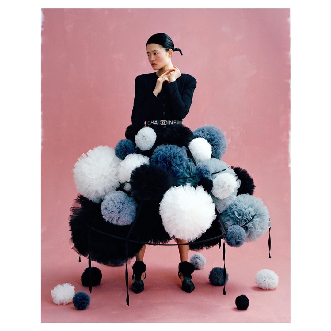 Asian woman in black blazer seated behind black metal structure covered in black, white, and grey pom poms against pink backdrop.