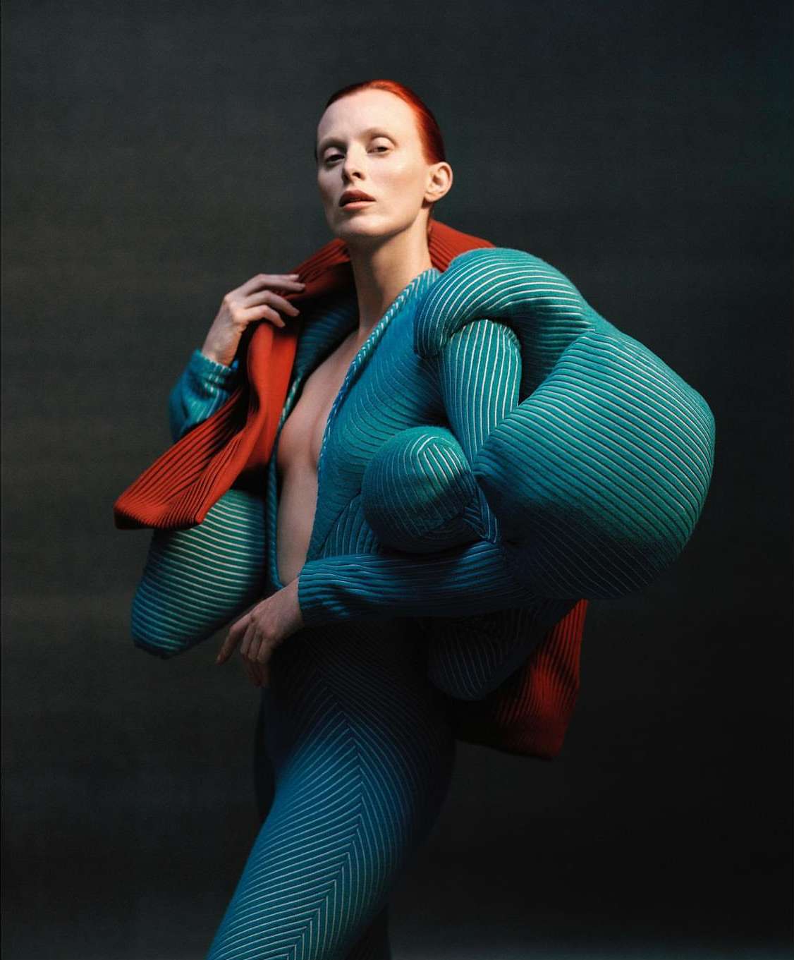Fashionable redhead woman in futuristic ribbed outfit with dramatic jacket poses in a studio with a dark background and neutral lighting.