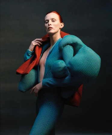 Fashionable redhead woman in futuristic ribbed outfit with dramatic jacket poses in a studio with a dark background and neutral lighting.