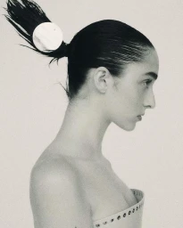 Profile portrait of a woman with dark hair in a sleek ponytail secured with a circular clip, wearing a structured strapless top against a light backdrop.