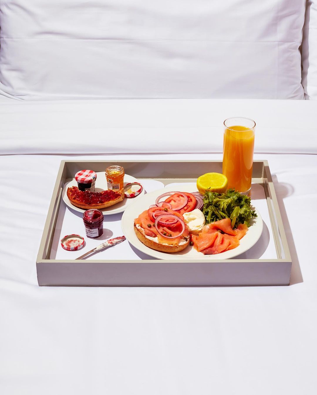 Breakfast in bed features bagels with salmon, tomatoes, onions, lettuce, and cream cheese, alongside orange juice and jam on a grey tray.