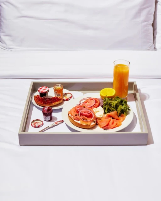 Breakfast in bed features bagels with salmon, tomatoes, onions, lettuce, and cream cheese, alongside orange juice and jam on a grey tray.