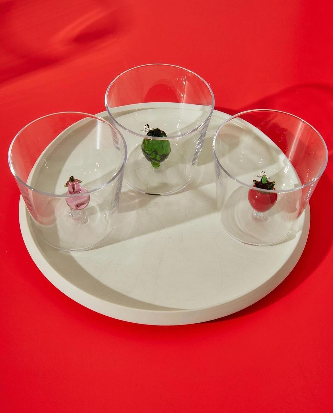 Three clear glasses sit on a round white tray against a red background, each with a different color fruit ornament inside the glass.