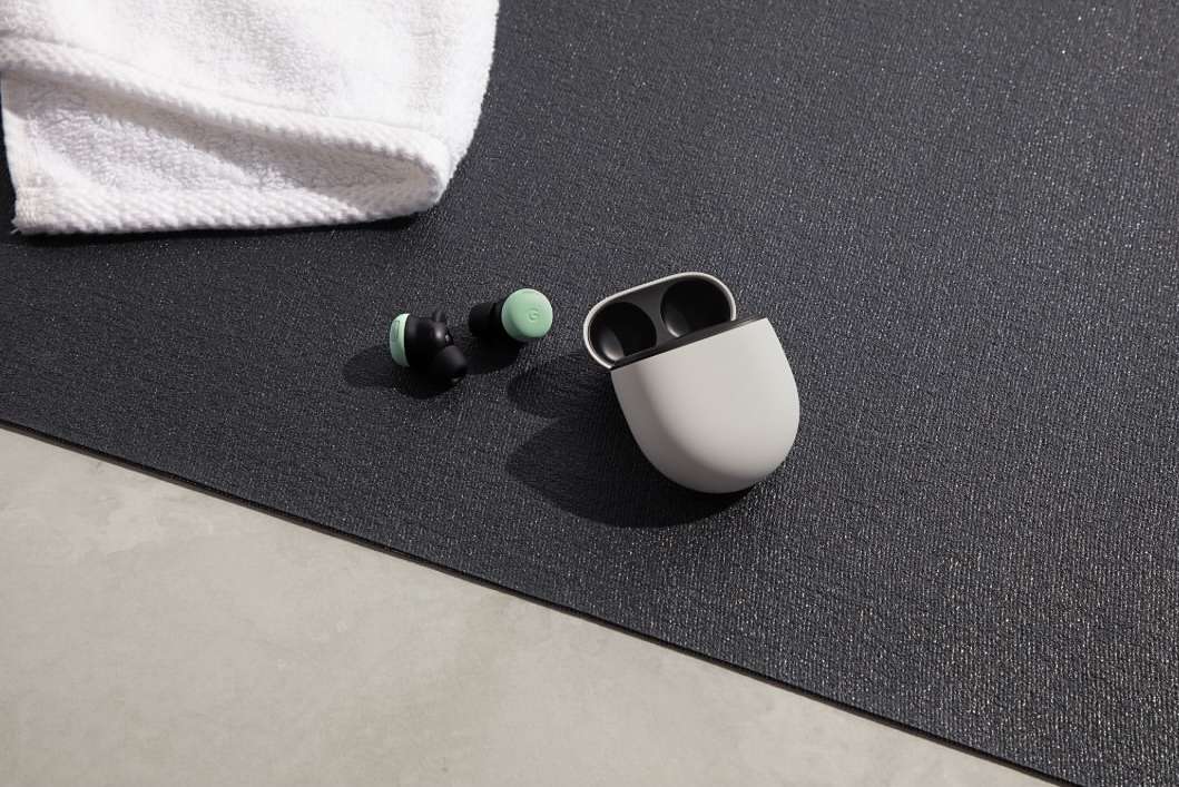 Close-up of wireless earbuds with a charging case and white towel on a textured grey fitness mat on a light grey floor.