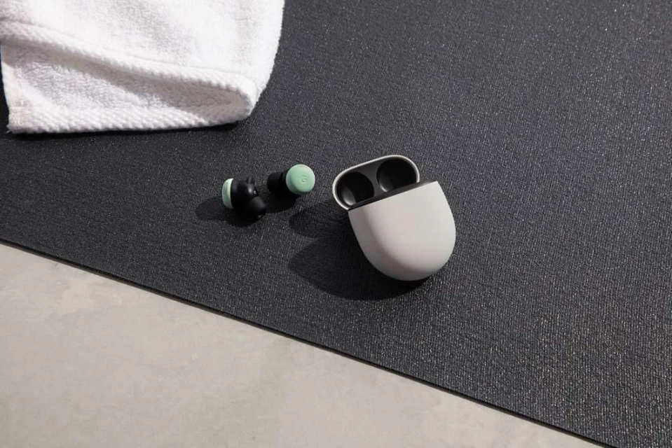 Close-up of wireless earbuds with a charging case and white towel on a textured grey fitness mat on a light grey floor.