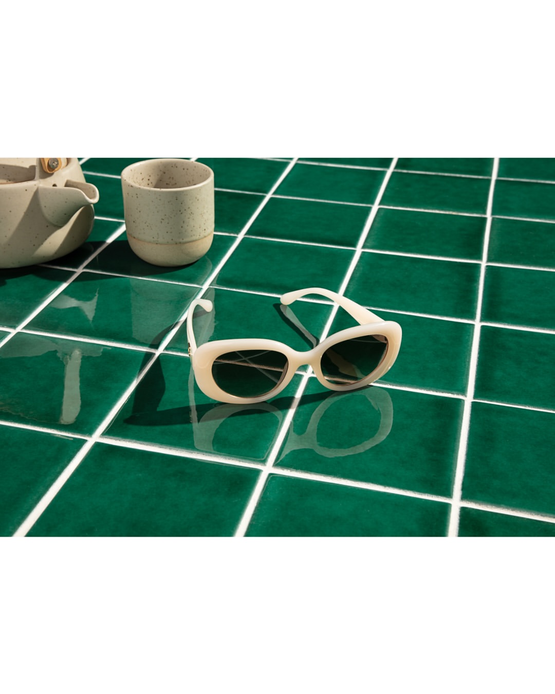 White sunglasses and a ceramic cup resting on a glossy green tiled tabletop.