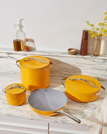 Still life of yellow cookware set including pots, pans, and lids on a marble countertop in a bright, minimalist kitchen setting.