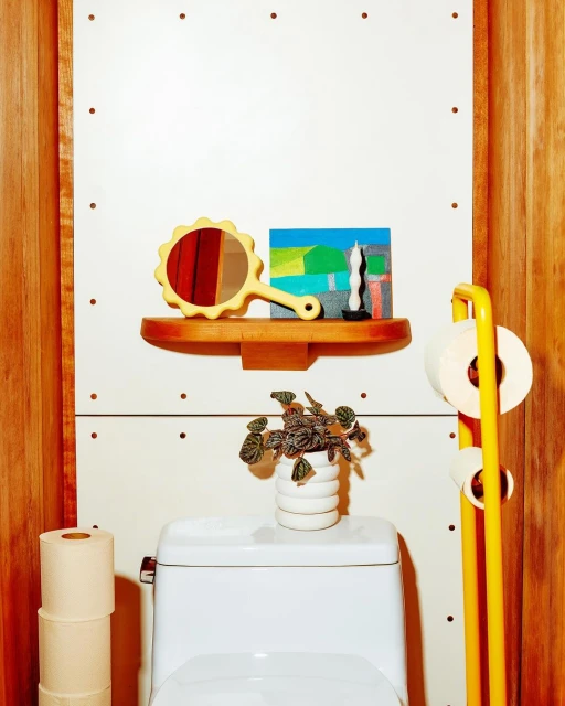 Bright, well-lit shot of a modern, minimalist bathroom featuring a white toilet, shelf decorations, a potted plant, and a yellow toilet paper holder.
