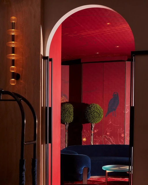 Interior view through an arched doorway with red walls, topiary trees, blue velvet sofa, table, and a decorative wall light fixture.