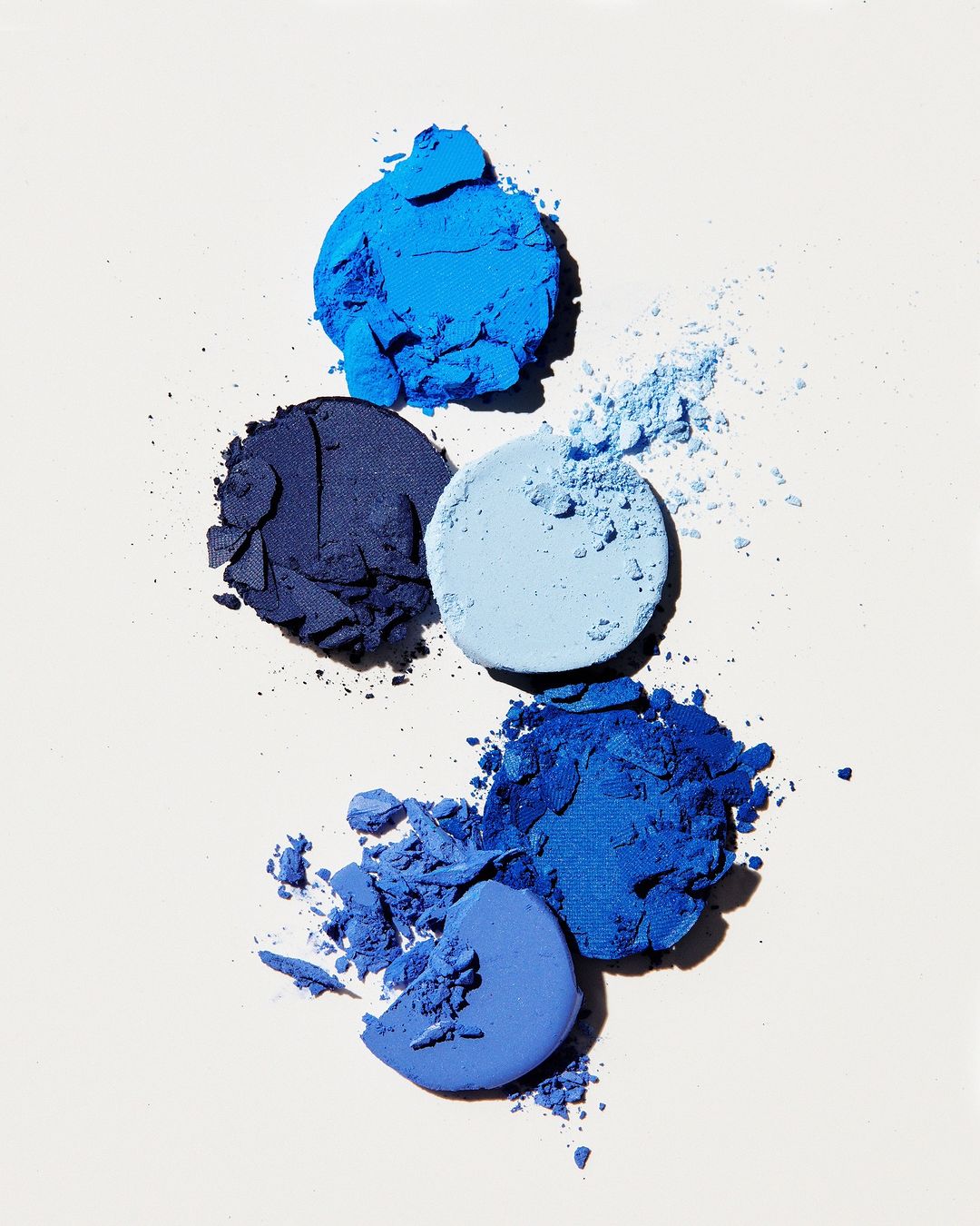Overhead shot of blue eyeshadows, crushed and intact, in a variety of blue tones, displayed against a pale background.