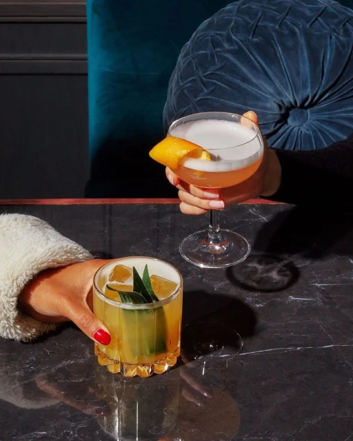 Two cocktails are presented on a marble table, one coupe glass garnished with an orange peel, and a rocks glass with a tropical leaf.
