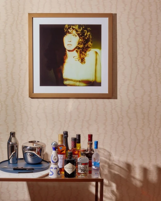 Bar cart with liquor bottles below a framed photo of a woman with curly hair against a patterned beige wall in an indoor setting.