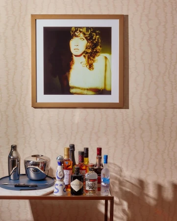 Bar cart with liquor bottles below a framed photo of a woman with curly hair against a patterned beige wall in an indoor setting.