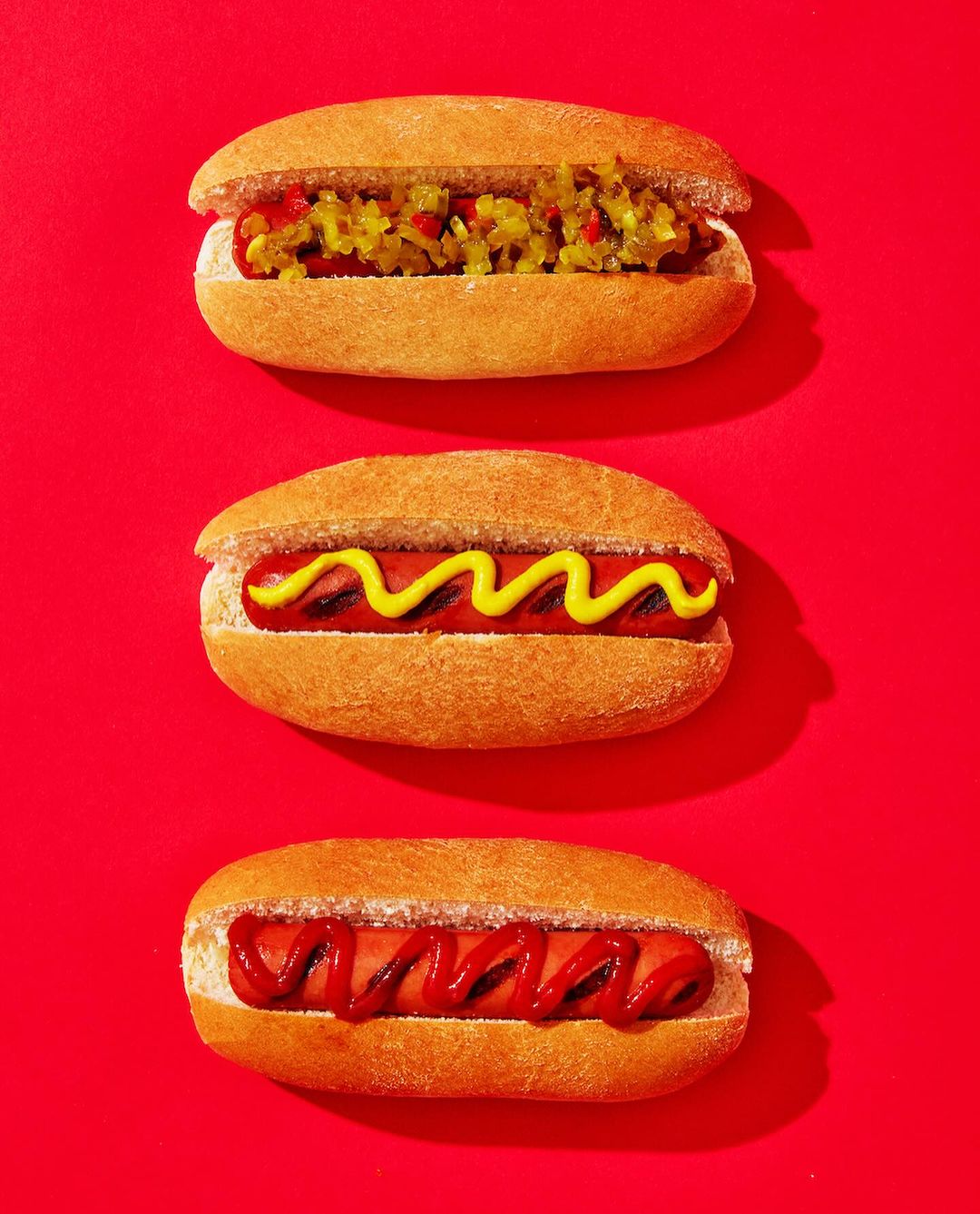 Overhead shot of three hot dogs in buns with different toppings: relish, mustard, and ketchup, arranged vertically on a red surface.
