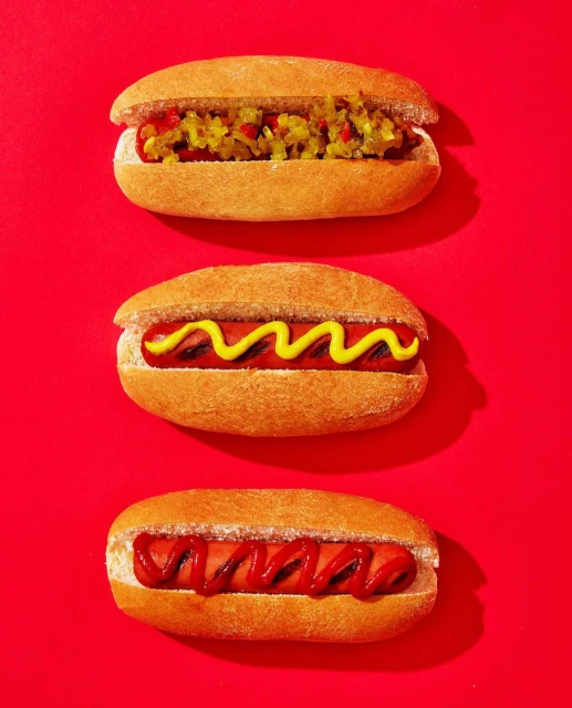 Overhead shot of three hot dogs in buns with different toppings: relish, mustard, and ketchup, arranged vertically on a red surface.