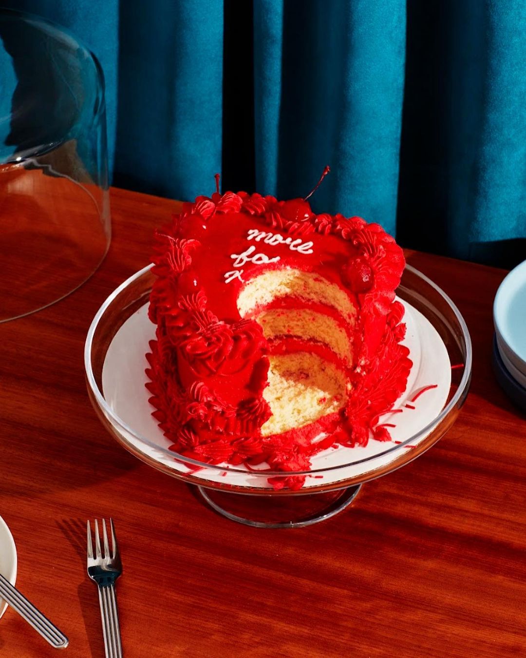 Red velvet cake with white frosting on a clear cake stand, set on a wooden table against a teal curtain background. Food photography.