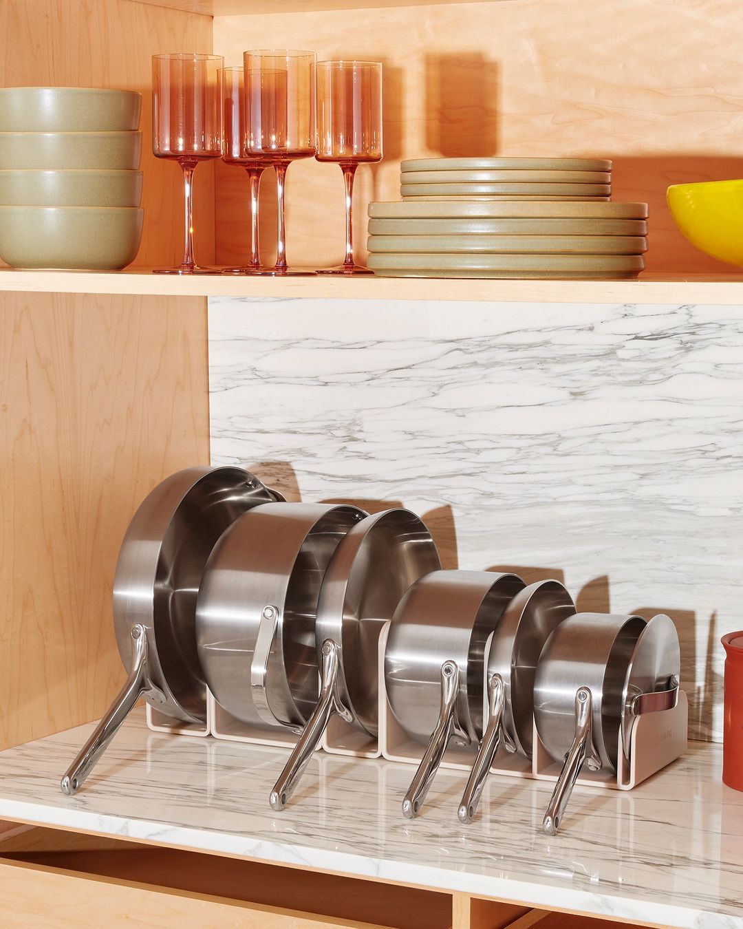 Organized collection of stainless steel pots and pans in a kitchen cabinet with matching tableware and pink stemmed glassware.