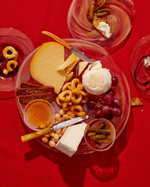 Overhead shot of a clear plate filled with assorted cheeses, crackers, grapes, nuts, and honey, set against a vibrant red background.
