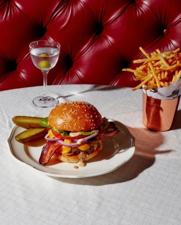 Gourmet burger with bacon, lettuce, tomato, onion, and cheese on a plate with pickles, served with fries and a martini.