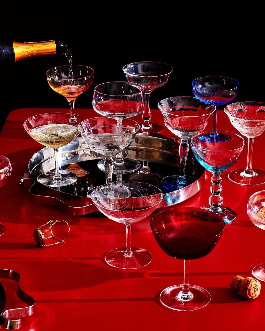 Champagne is poured into a coupe glass in a still life of cocktail glasses, champagne bottle, and corks on a red surface.