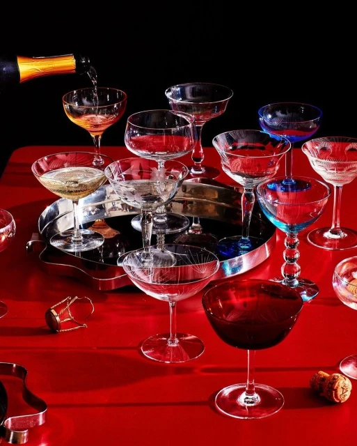 Champagne is poured into a coupe glass in a still life of cocktail glasses, champagne bottle, and corks on a red surface.