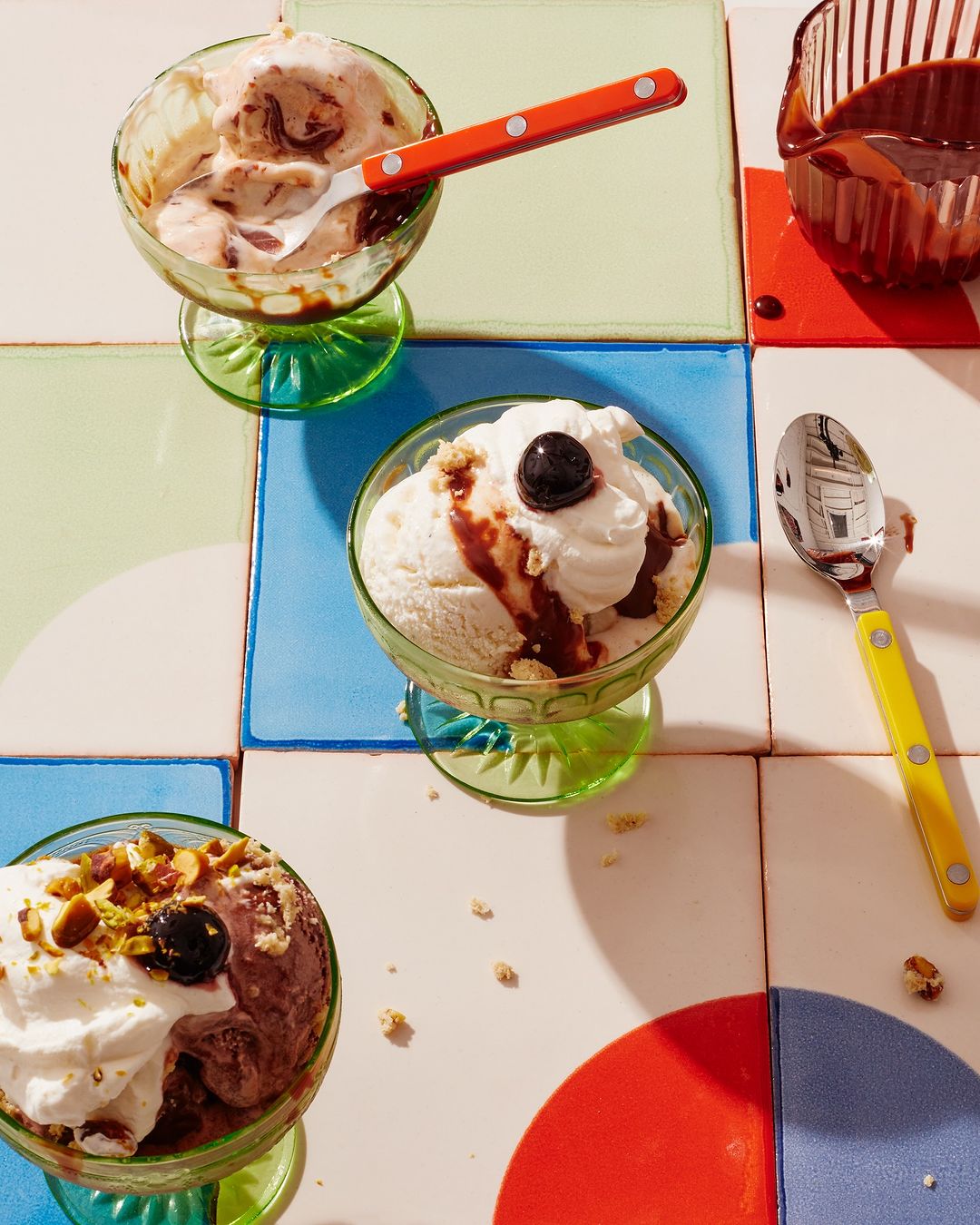 Three glass dessert bowls filled with ice cream, whipped cream, toppings, and spoons arranged on a colorful tiled surface.