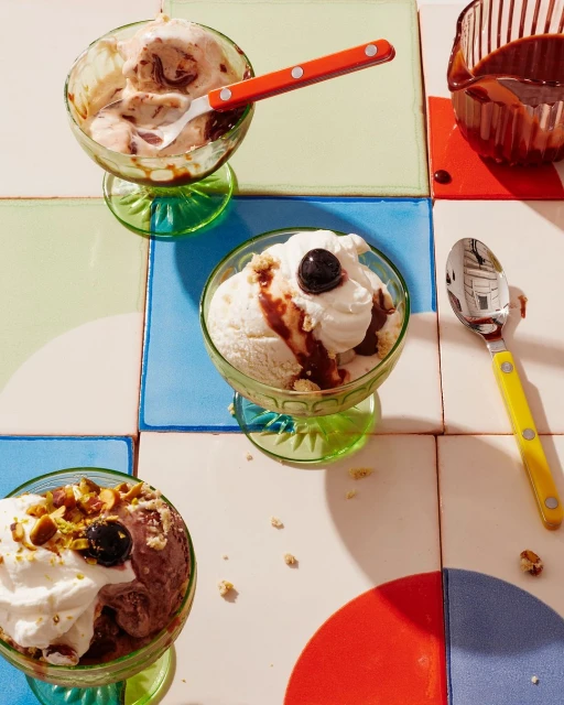 Three glass dessert bowls filled with ice cream, whipped cream, toppings, and spoons arranged on a colorful tiled surface.