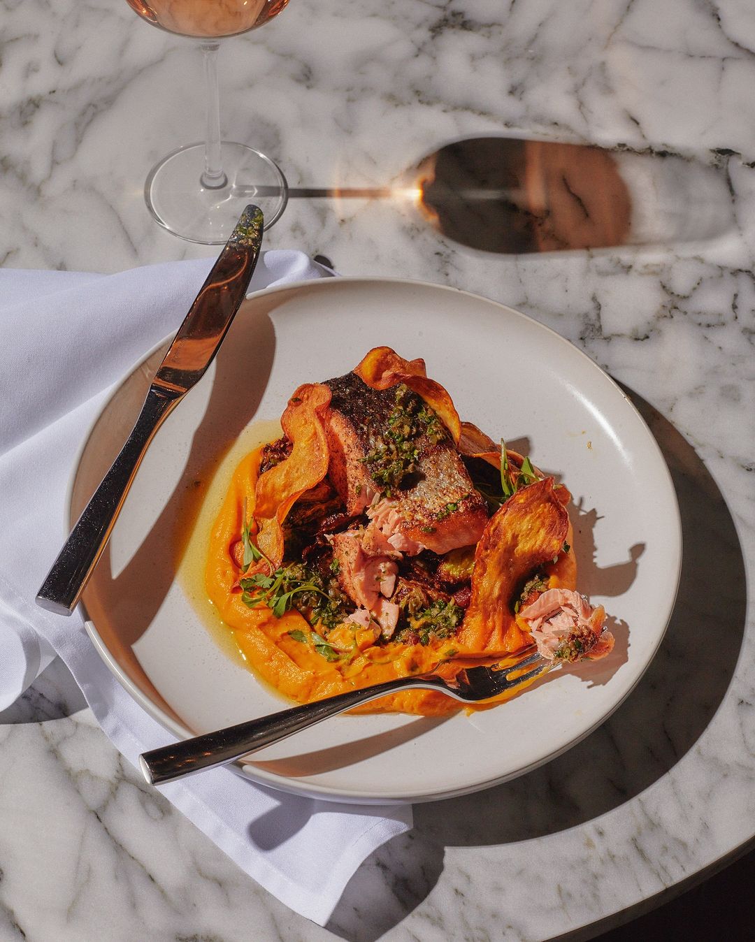 Overhead shot of a salmon dish with orange puree, herbs, and a wine glass on a marble table, showcasing culinary art and food photography.