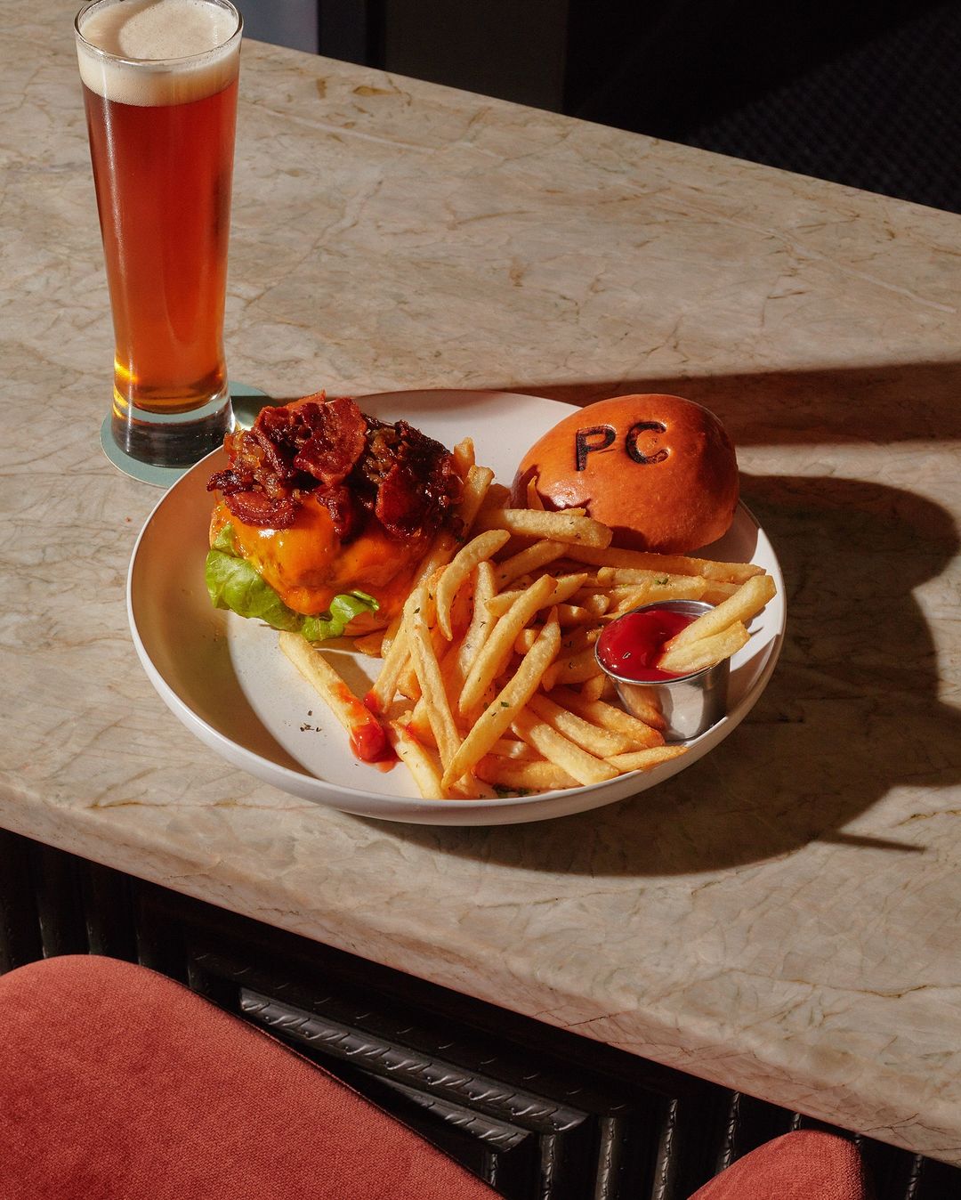 Overhead shot of a hamburger with bacon, fries, and beer on a white plate and light marble table in soft, natural lighting.